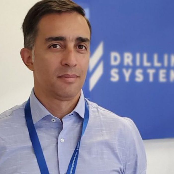 A man wearing a light blue shirt and blue lanyard stands in front of a blue sign that reads "DRILLING SYSTEMS.