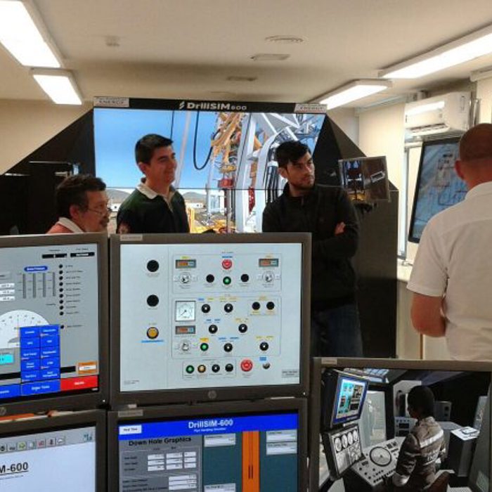 A group of people stand in a room with various monitors and control panels, attentively listening to a man giving a presentation in front of a large screen.