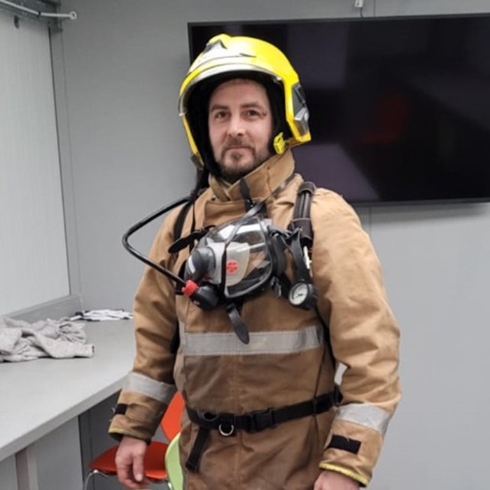 A person wearing firefighting gear, including a yellow helmet and breathing apparatus, stands in a room with a desk and a large screen on the wall.
