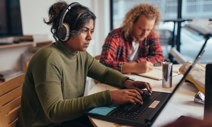 A person with headphones works on a laptop at a table, focused on training modules, while another person writes in the background.