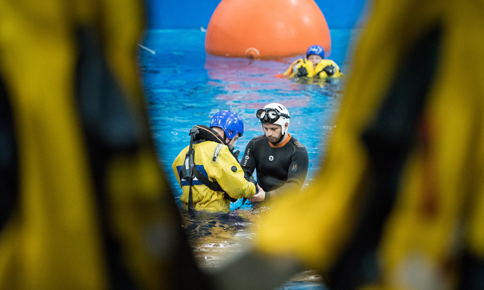 Two individuals in water activity gear are seen in the center of a pool, surrounded by others in yellow suits, with a large orange buoy in the background.