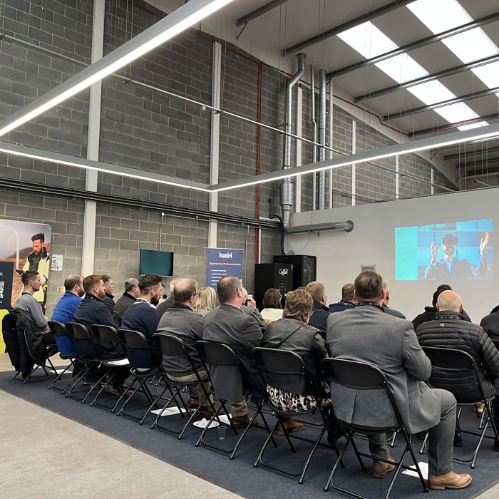 A group of people in rows attentively engages in a training session, facing a speaker and a large screen, all within the sleek ambiance of a modern industrial-looking conference room.