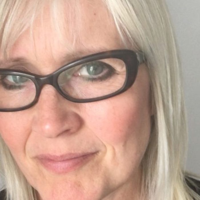 A person with straight, light-colored hair and green eyes is wearing dark-framed glasses and looking directly at the camera against a plain background.