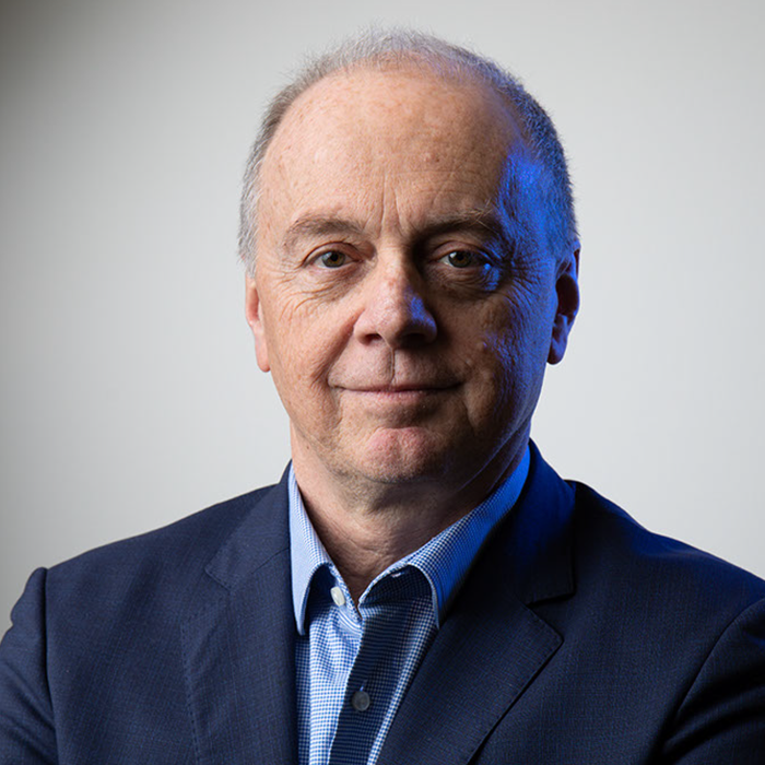 A middle-aged man with a receding hairline, wearing a blue suit jacket and a blue shirt, stands against a plain background with a neutral expression.