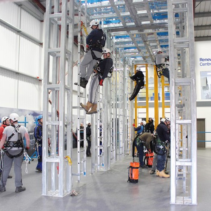 People in safety gear and helmets practice climbing on a metal structure in an indoor training center labeled as the Renewable Energy Centre of Excellence.