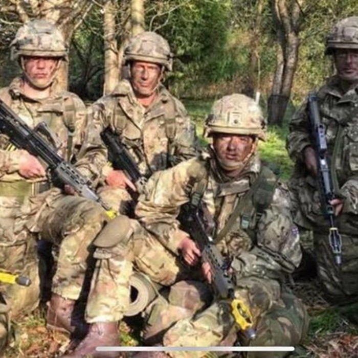 A group of soldiers in camouflage uniforms and helmets pose for a photo in a forested area, holding rifles with yellow attachments.