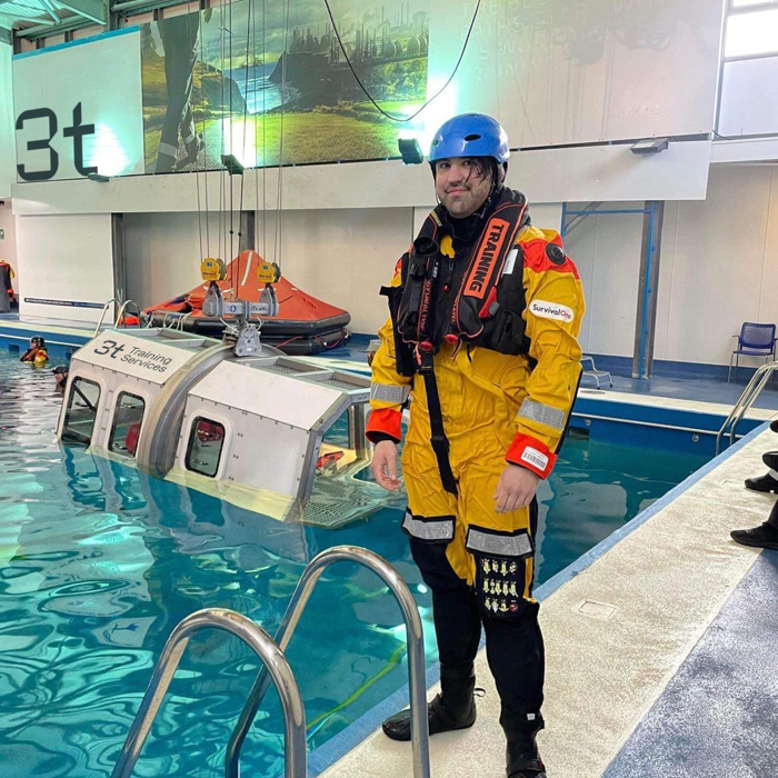 A person in water survival gear stands by a training pool, where a mock helicopter is partially submerged for a realistic training scenario.