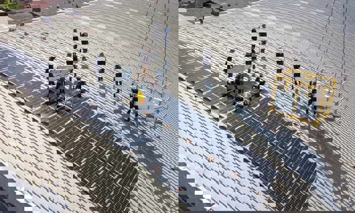 Workers installing solar panels on a large, ribbed metal rooftop, using a crane to lift equipment.