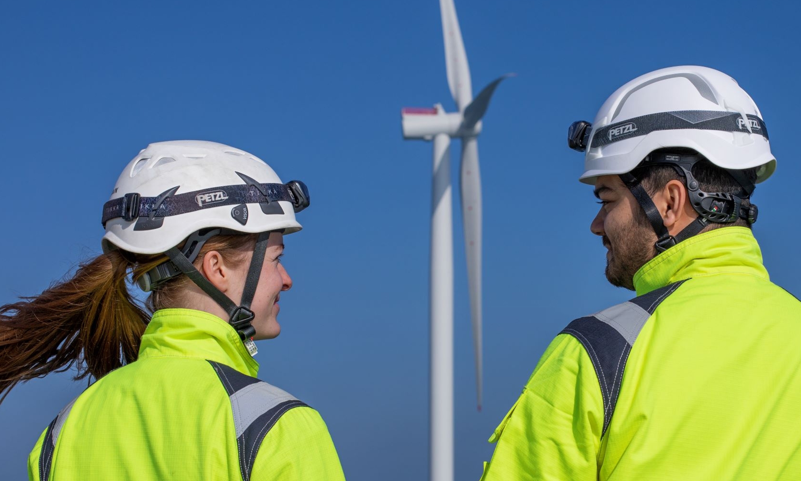 Two workers in high-visibility jackets and helmets, recently engaged in training, stand near an offshore wind turbine against a clear blue sky.
