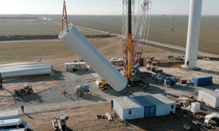 A crane is lifting a large cylindrical section of a wind turbine tower at a construction site; various construction vehicles, equipment, and materials are scattered around.