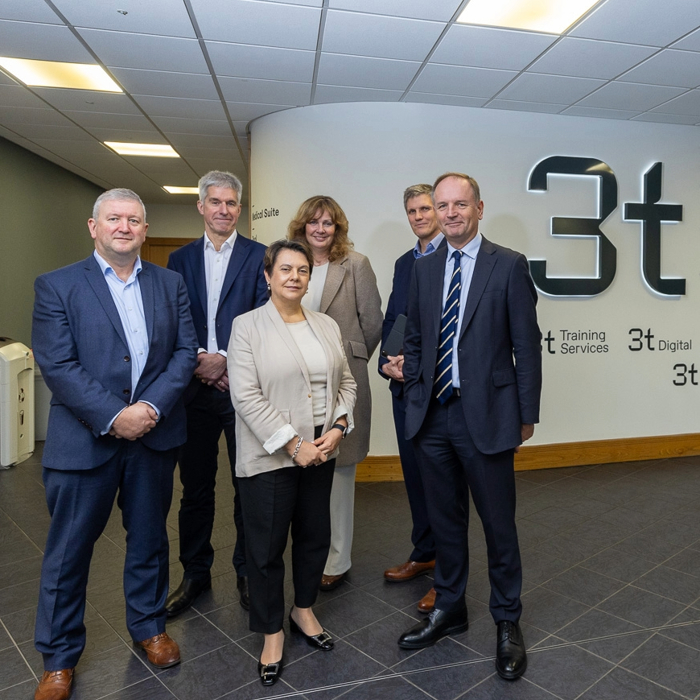 Six people in business attire stand in a tiled hallway next to a wall with "3t" logos.