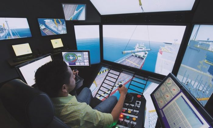 A person operates a complex maritime simulator with multiple screens, showing a ship being loaded. Various control panels and monitors are visible in the simulator room.