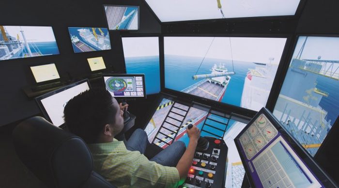 A person operates a complex maritime simulator with multiple screens, showing a ship being loaded. Various control panels and monitors are visible in the simulator room.