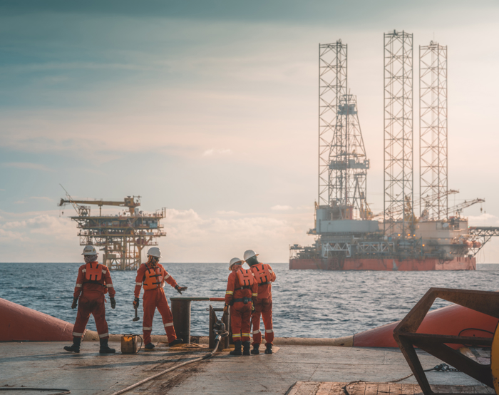 Quatro trabalhadores com equipamento de segurança estão numa plataforma offshore, aperfeiçoando as suas competências através de formação prática enquanto executam tarefas, com duas plataformas petrolíferas visíveis ao fundo sobre o mar.