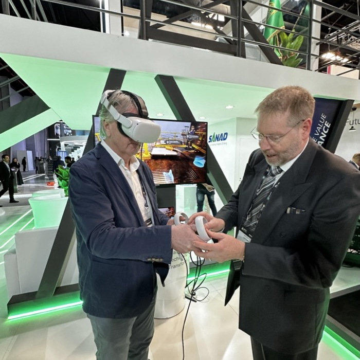 Two men at an exhibition; one wearing a VR headset and holding a controller, immersed in a training simulation, while the other assists. The booth is bathed in green lighting with a screen displaying dynamic visuals in the background.