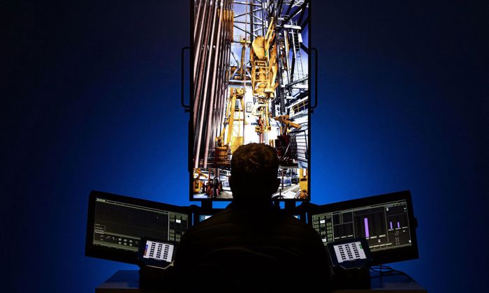 A person sits at a control station with multiple monitors displaying industrial machinery, suggesting an operation or monitoring of equipment.