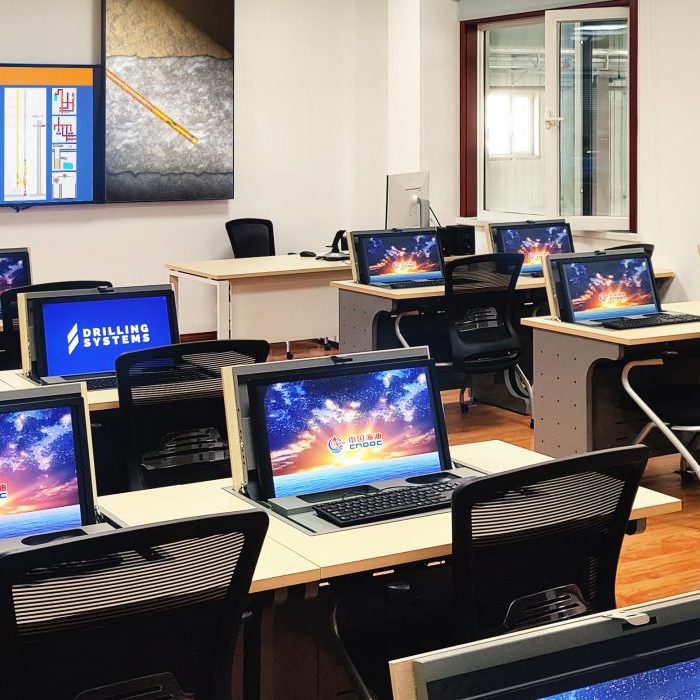 A computer lab with multiple rows of desks, each equipped with a desktop computer featuring brightly colored screensavers. The room includes a large display screen at the front.