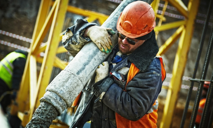 Construction worker wearing an orange helmet and safety gear, holding a large concrete hose, with a construction site background.