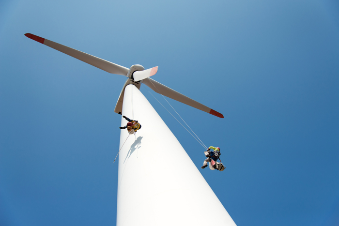 Two workers, supported by ropes, carry out maintenance on a large white wind turbine against a clear blue sky. Their expertise is the result of funded training programs designed to ensure safety and efficiency in this high-risk environment.