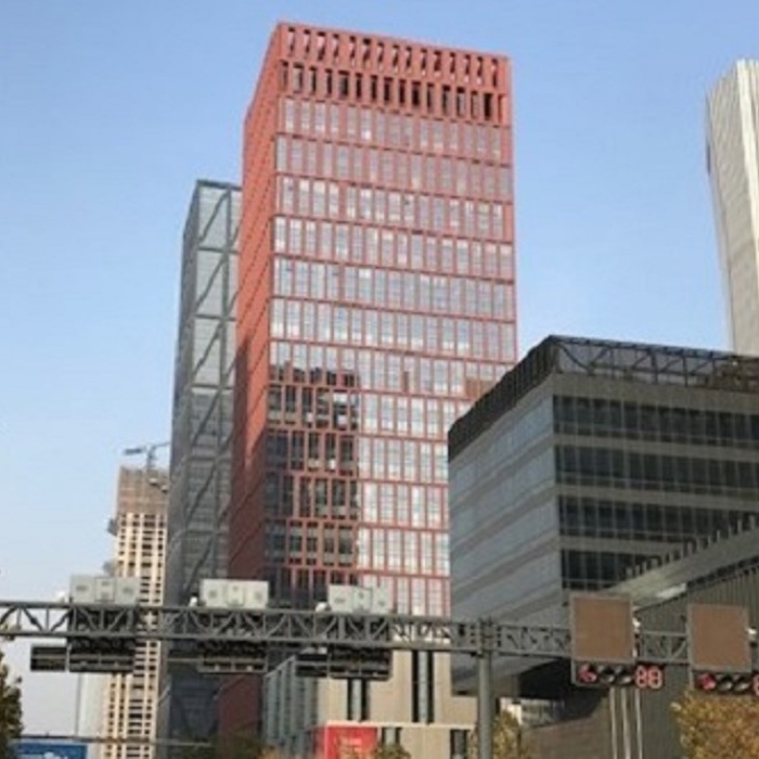 A cityscape featuring a red high-rise building flanked by other tall, modern buildings, with a traffic signal gantry visible in the foreground.