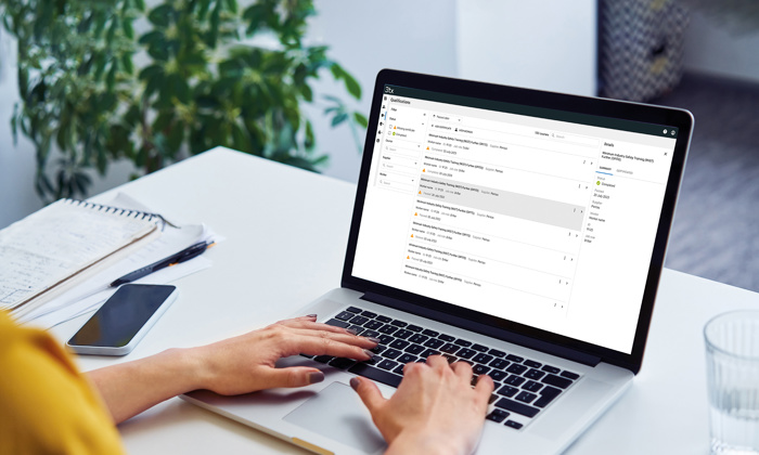 Pessoa a escrever num computador portátil numa secretária com uma caixa de correio eletrónico aberta no ecrã, junto a um bloco de notas, uma caneta, um smartphone e um copo de água. Uma planta em segundo plano.