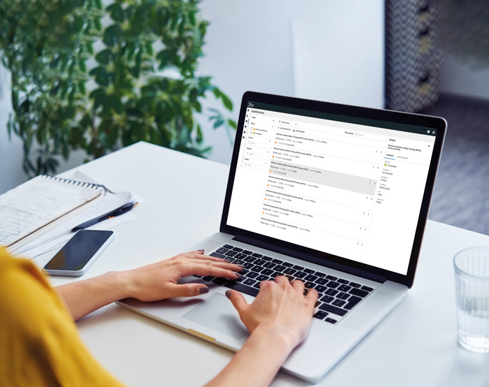 Pessoa a escrever num computador portátil numa secretária com uma caixa de correio eletrónico aberta no ecrã, junto a um bloco de notas, uma caneta, um smartphone e um copo de água. Uma planta em segundo plano.
