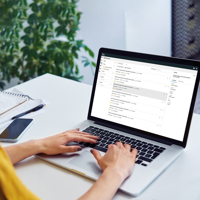 Pessoa a escrever num computador portátil numa secretária com uma caixa de correio eletrónico aberta no ecrã, junto a um bloco de notas, uma caneta, um smartphone e um copo de água. Uma planta em segundo plano.