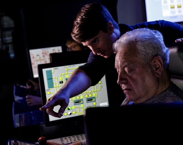 An older man operates controls while a younger man points at something on a screen in a control room setting with multiple monitors.