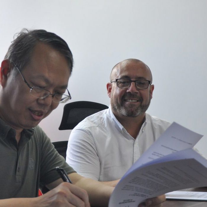 Two men are seated at a table with documents in front of them. One man is signing a paper, while the other smiles. A red banner with text in a foreign language is displayed in the background.