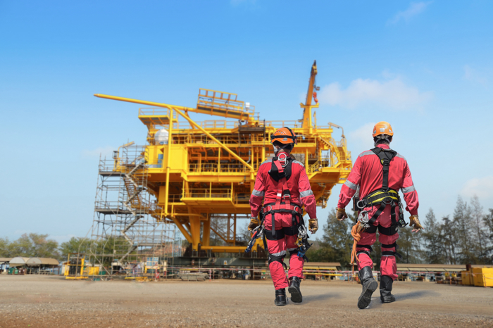 Two workers in red protective gear walk towards a large yellow oil rig under a clear blue sky, exemplifying the precision and skill gained from rigorous training.