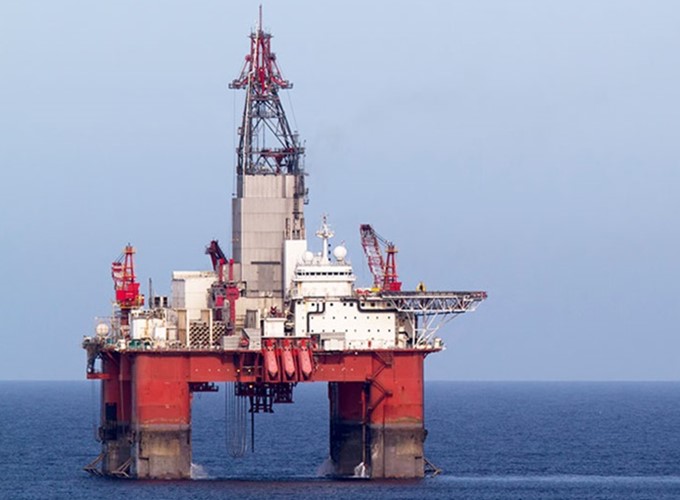 An offshore oil rig stationed in the ocean, with red and white machinery and equipment visible against a clear sky.