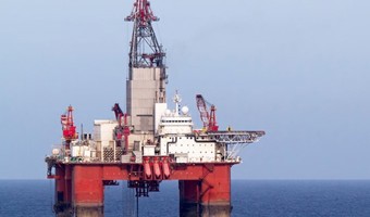 An offshore oil rig stationed in the ocean, with red and white machinery and equipment visible against a clear sky.