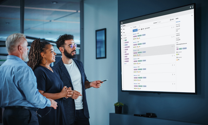 Three people stand in an office, looking at a large screen displaying a training schedule. One person is pointing at the screen.
