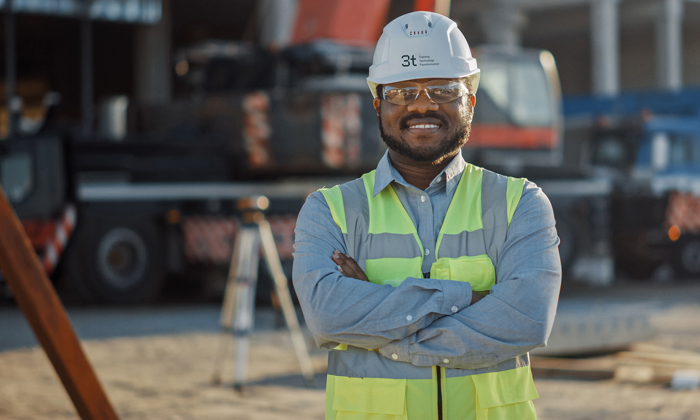 A construction worker in a safety vest and hard hat stands with arms crossed at a construction site.