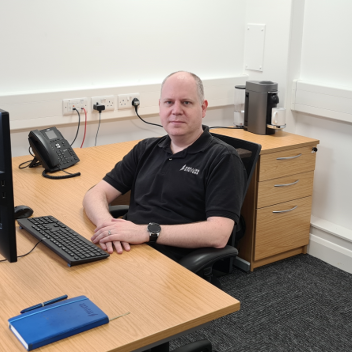 A person seated at a wooden desk with two monitors, keyboard, phone, and notebook, in an office. The person is wearing a black shirt and looking at the camera.