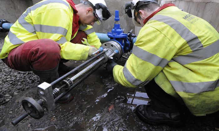 Two workers in yellow safety jackets and helmets are fixing a pipe valve outdoors on a muddy ground.