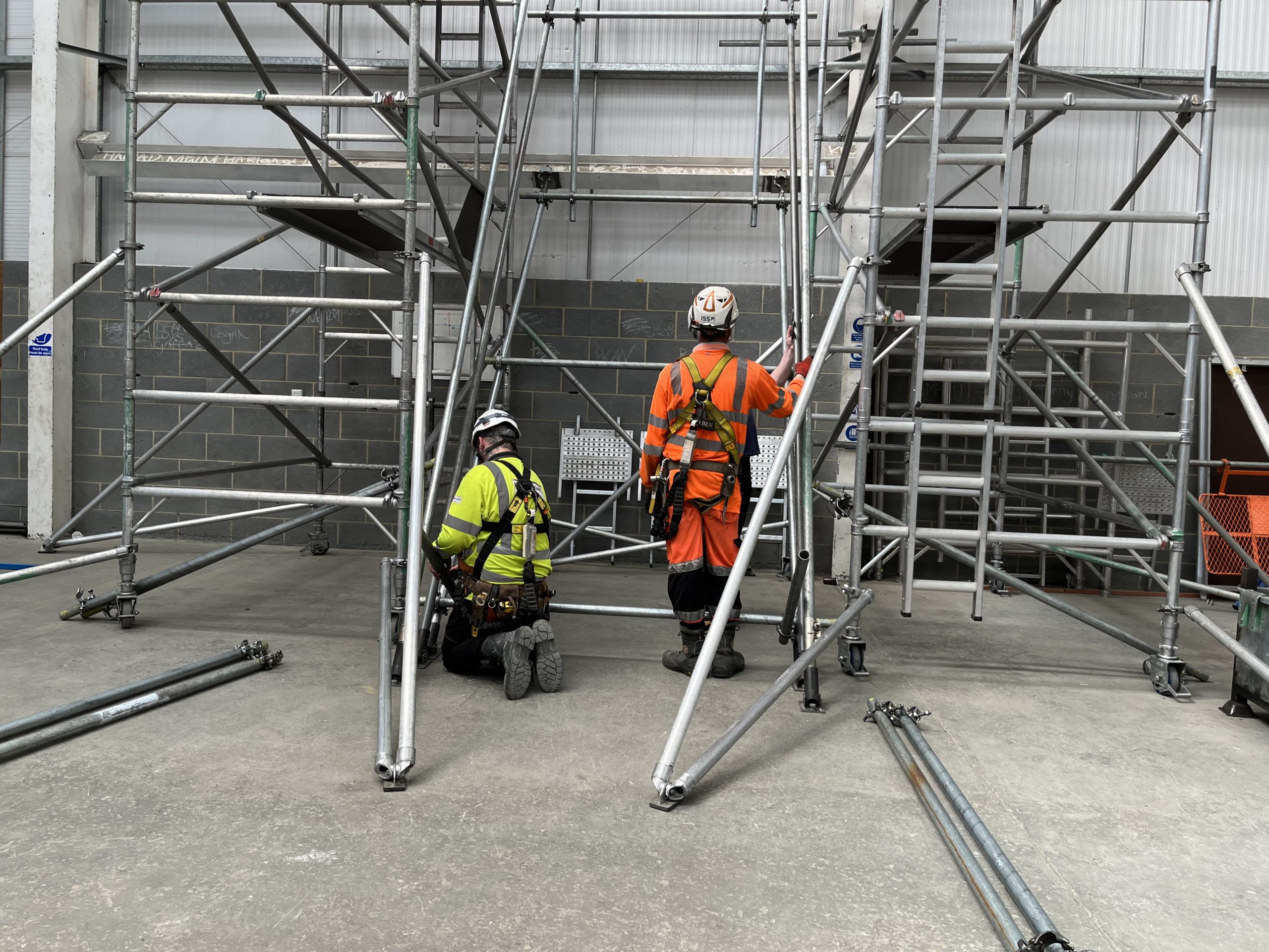 Two workers in safety gear carefully assemble scaffolding in an industrial setting, demonstrating precise training and expertise.