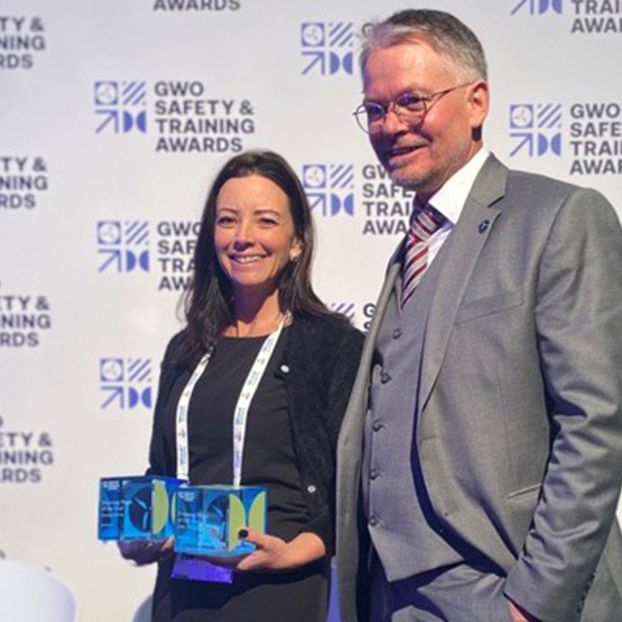 Two people stand smiling. The woman holds two awards, and a man stands beside her, both in business attire. Background has "GWO Safety and Training Awards" repeated.
