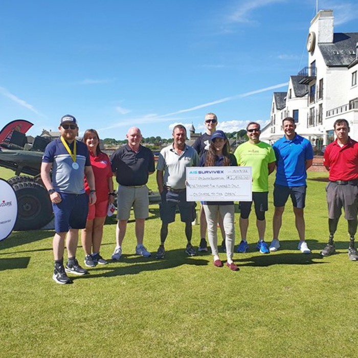 A group of people stand on a golf course holding a large check. Some participants are wearing casual sportswear, and one person in the group has a prosthetic leg.