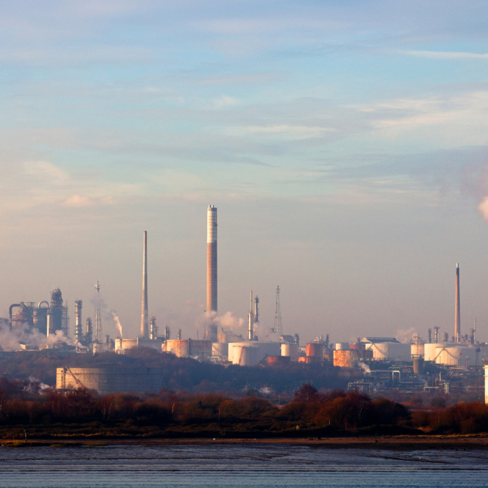 An industrial refinery landscape sprawls beneath a blue sky with scattered clouds, its chimneys puffing smoke like sentinels in training, synchronized and steadfast.