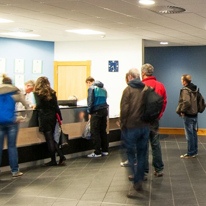 People are standing in line at a reception counter in a modern office lobby. Some are engaging with the staff, while others await their turn. The lobby has tiled flooring and blue accent walls.