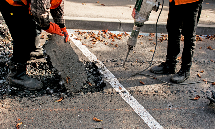 Two workers in safety gear, adept from rigorous training, break and remove a section of asphalt on a road using a jackhammer.