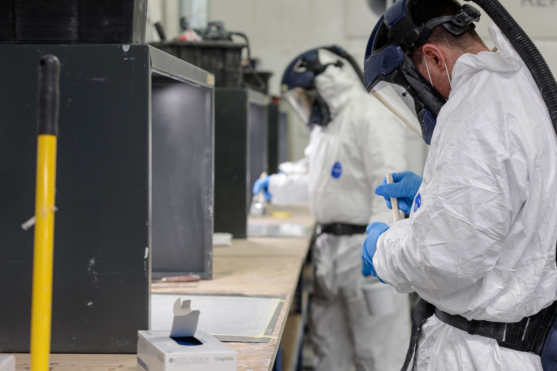 Two people in protective suits and helmets engage in a training session at the lab bench, carefully handling materials with gloves. A box and a yellow pole are visible in the foreground.