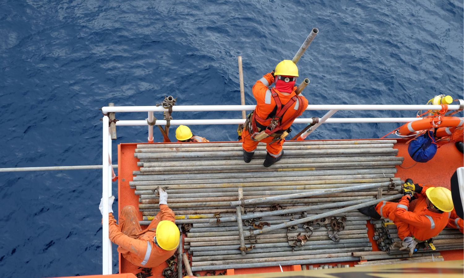Trabalhadores com equipamento de segurança cor de laranja e capacetes amarelos manuseiam tubos de metal no convés de uma plataforma offshore, tendo como pano de fundo o oceano.