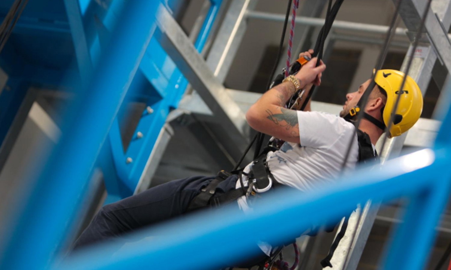 A person in a yellow helmet and harness is undergoing training as they ascend ropes in an industrial setting, with blue metal beams providing a striking backdrop.