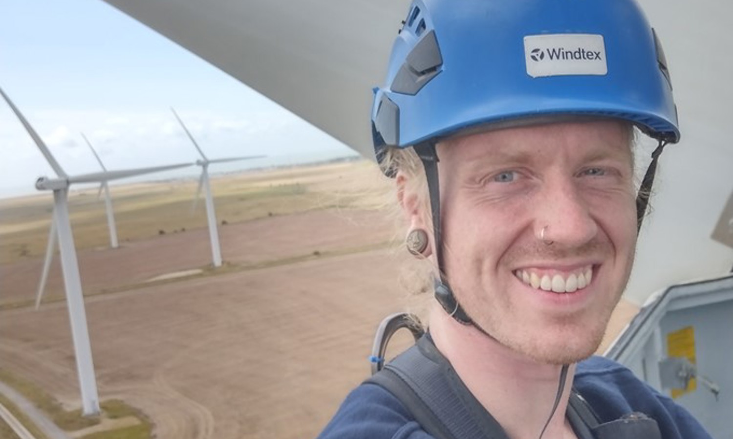 A person wearing a blue helmet labeled "Windtex" takes a selfie from a high vantage point with wind turbines and wide fields visible in the background.