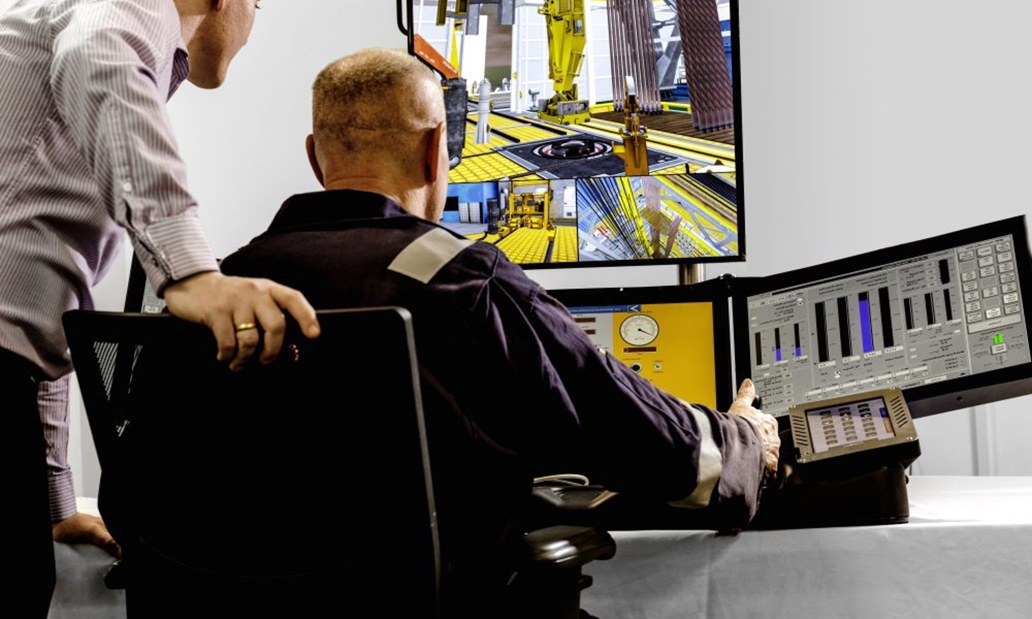 Two men in a control room monitor multiple screens displaying industrial machinery and data charts. One man sits while the other leans over, observing the screens.