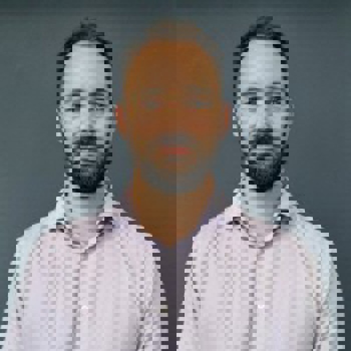 Man with short hair and a beard, wearing a light-colored dress shirt, stands confidently as if ready for training, set against a plain dark background.