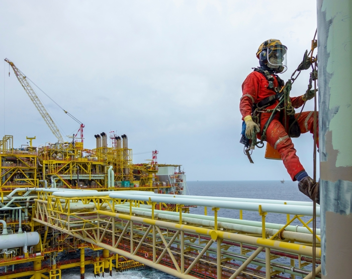 A worker in safety gear, demonstrating expert training, is suspended on ropes inspecting a vertical structure on an offshore oil rig, with ocean and platform machinery visible in the background.
