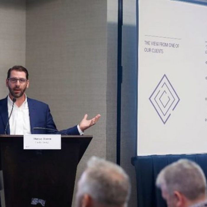 A man in a suit and glasses stands at a podium with outstretched arms, addressing an audience. A presentation board with bullet points and a logo is visible in the background.